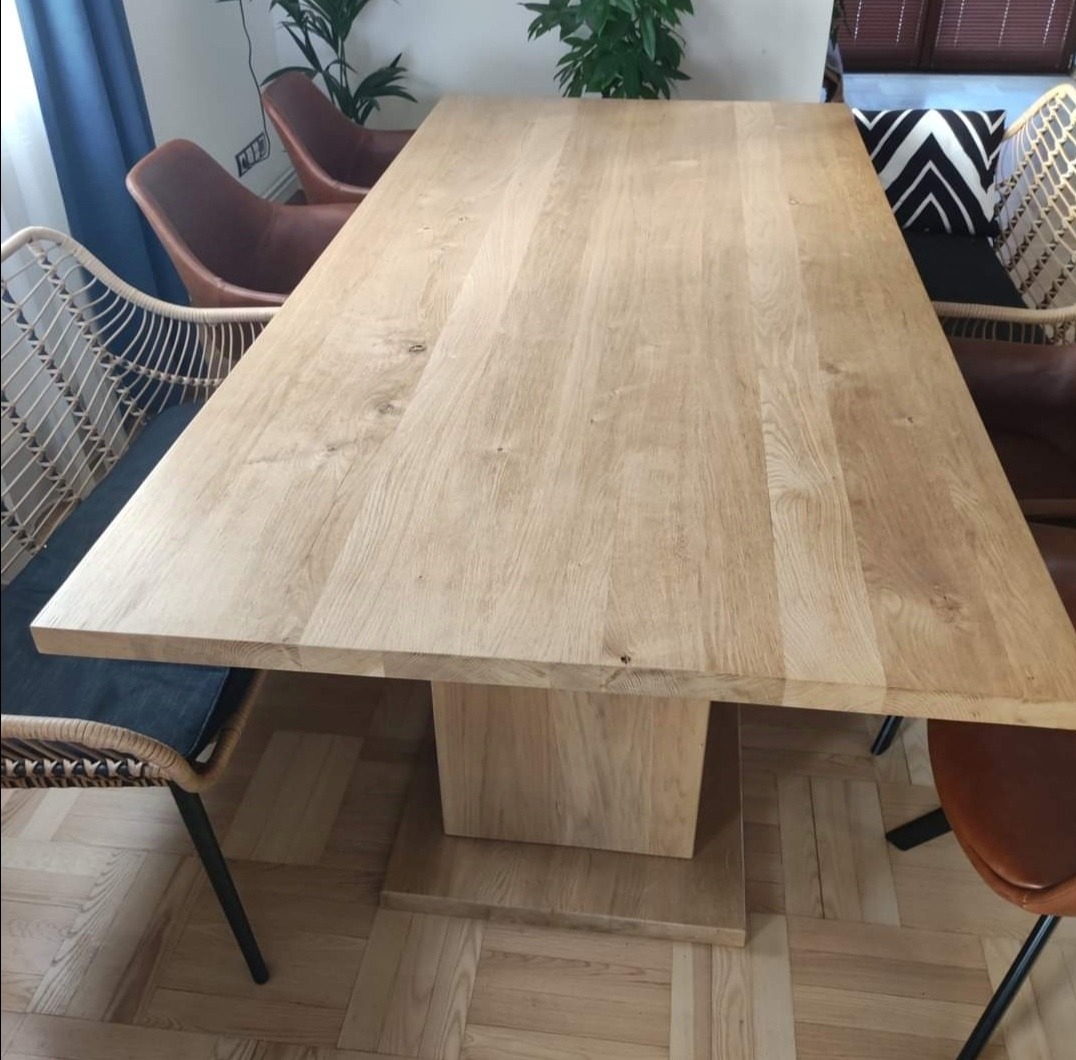 Duży, jasny stół z drewna z prostokątnym blatem i masywną nogą, otoczony krzesłami w różnym stylu, na parkiecie w jodełkę, Piskorowice.