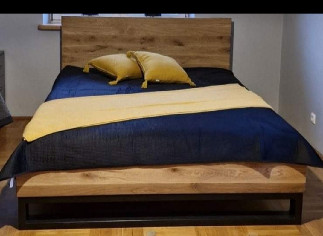 Łóżko z drewnianym wezgłowiem i ramą na metalowych nogach, przykryte granatową narzutą i żółtymi poduszkami, w sypialni.