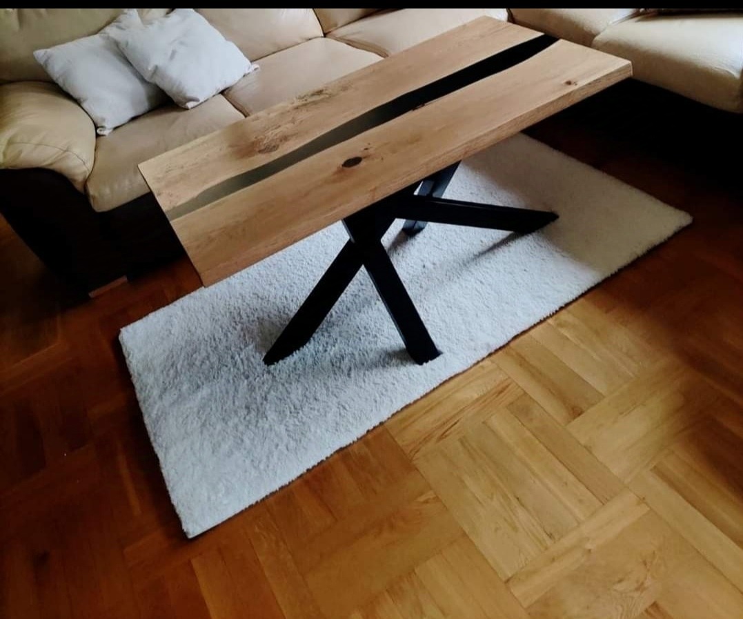 Stolik kawowy z blatem z jasnego drewna z czarną żywicą epoksydową wzdłuż środka, na czarnych, metalowych nogach w kształcie litery X, na białym dywaniku typu shaggy, na parkiecie.