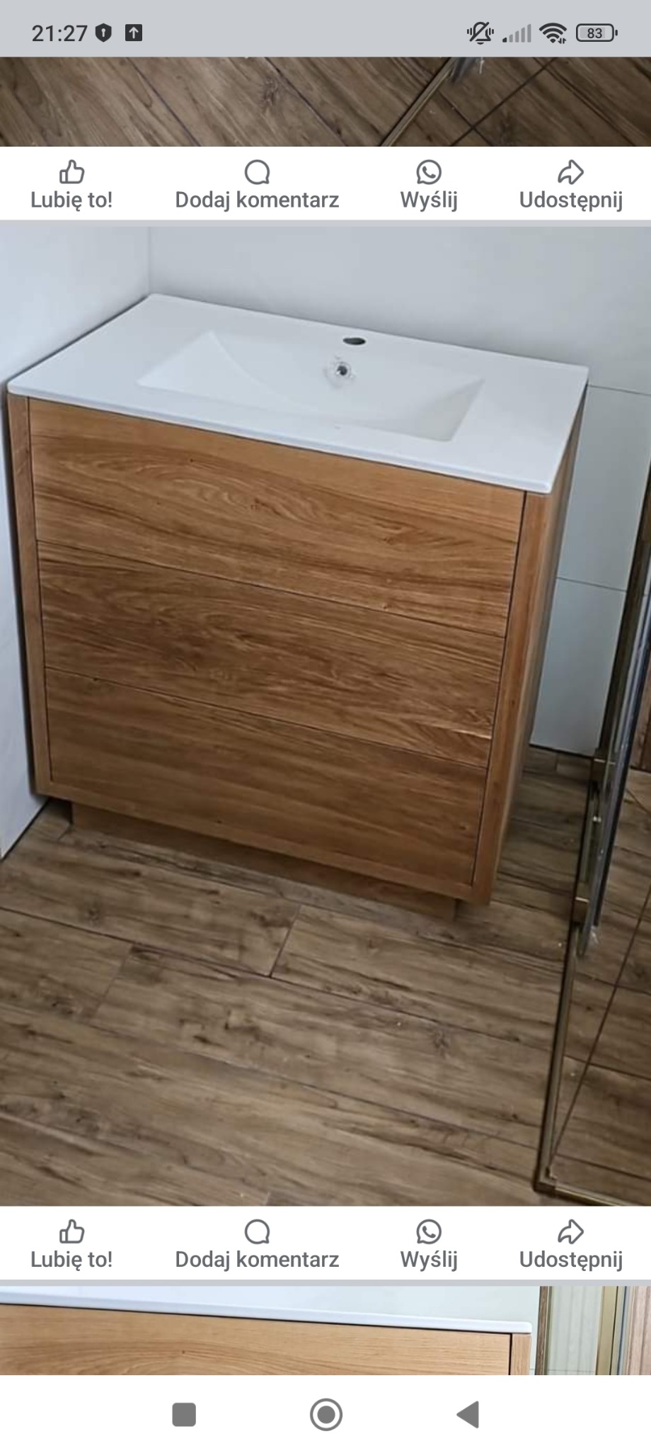 Szafka łazienkowa z drewnianym frontem i białą umywalką nablatową, widoczna na tle podłogi imitującej drewno i fragmentu kabiny prysznicowej.