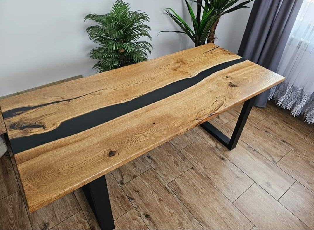 Blat stołu z drewna z żywicą epoksydową w kolorze czarnym, osadzony na metalowych nogach w kształcie trapezu, na tle roślin doniczkowych i podłogi z paneli imitujących drewno.