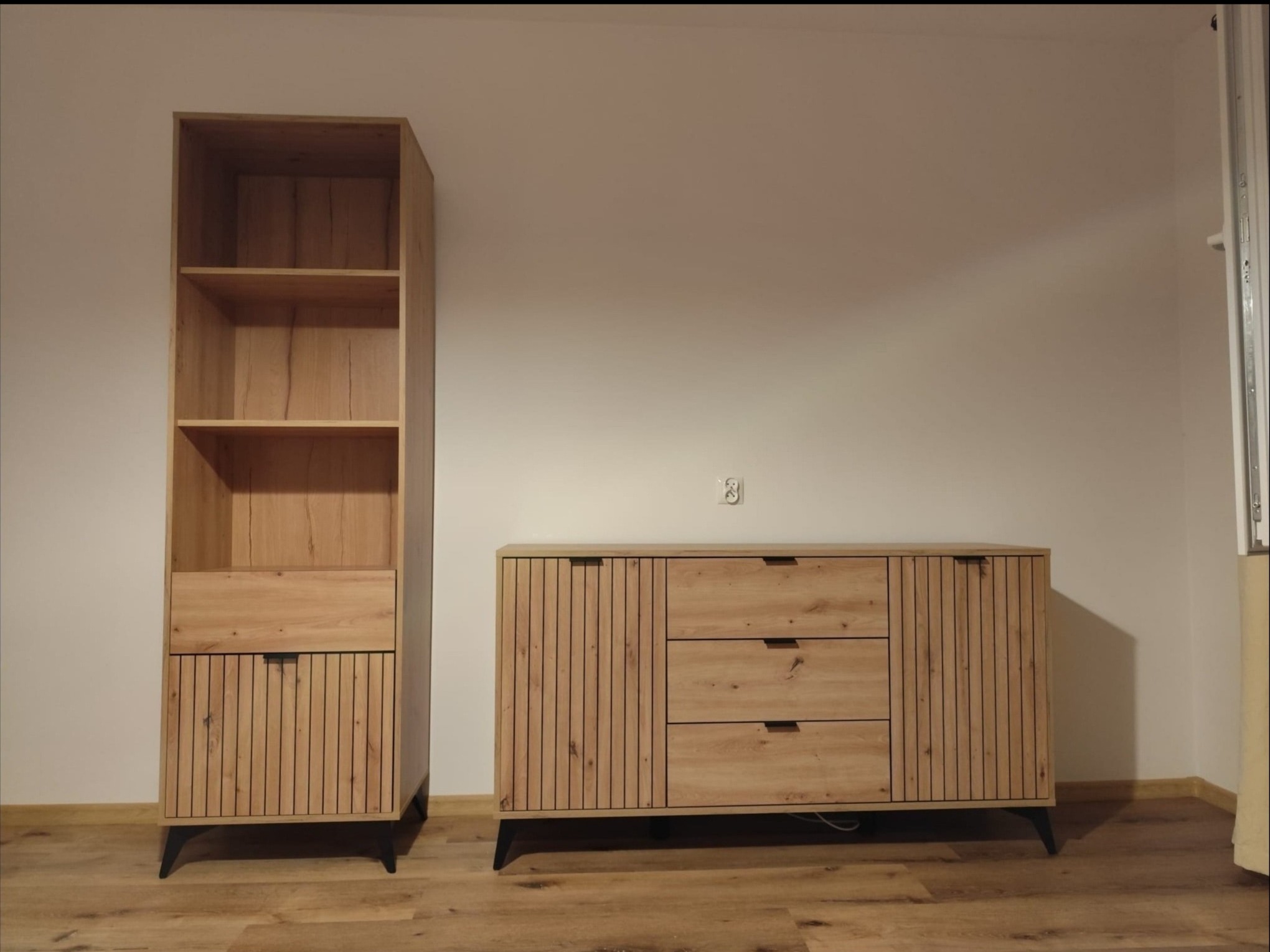 Zestaw mebli w stylu skandynawskim: wysoki regał z półkami i komoda z trzema szufladami, wykonane z drewna w jasnym odcieniu dębu, na czarnych nóżkach, ustawione na drewnianej podłodze.