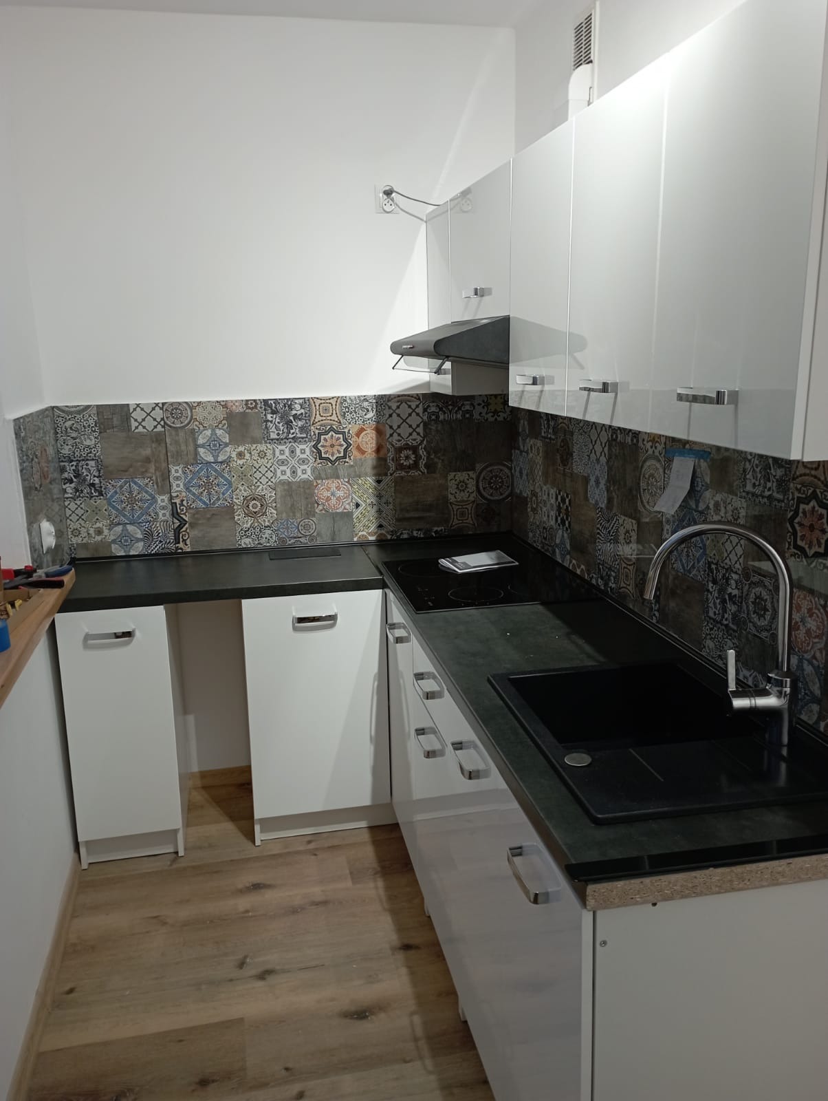 Nowoczesna kuchnia z białymi szafkami, czarnym blatem i dekoracyjnymi płytkami ściennymi z motywami patchworku, widok na zlewozmywak i płytę indukcyjną.