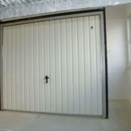 Zamknięta, segmentowa brama garażowa w kolorze białym z czarnym uchwytem w kształcie krzyża, widoczna z perspektywy wnętrza garażu z szarą podłogą i oknem po prawej stronie.