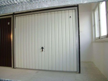 Zamknięta, segmentowa brama garażowa w kolorze białym z czarnym uchwytem w kształcie krzyża, widoczna z perspektywy wnętrza garażu z szarą podłogą i oknem po prawej stronie.