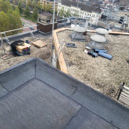 Dachwid - Widok z góry na płaski dach pokryty papą i drobnym żwirem, z elementami wentylacyjnymi, paletami i narzędziami, w tle zabudowa miejska.