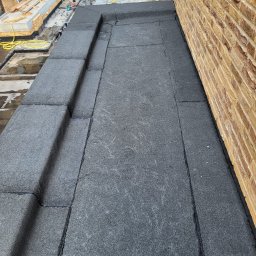 Dachwid - Nowa, płaska powierzchnia dachu pokryta papą termozgrzewalną, z widocznymi pracownikami w tle i żółtym wężem, przylega do ceglanej ściany budynku.