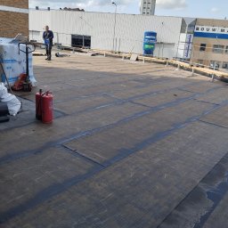 Dachwid - Płaski dach pokryty papą termozgrzewalną, widoczne zgrzewy, butle z gazem, paleta z materiałami budowlanymi i pracownik w tle. Częściowo zakończone prace.