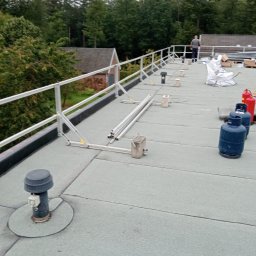 Dachwid - Płaski dach pokryty papą termozgrzewalną z zamontowaną aluminiową barierką ochronną, widoczne butle gazowe, elementy wentylacyjne i pracownik w oddali.