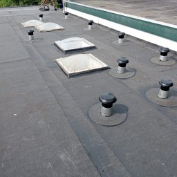 Dachwid - Płaski dach pokryty papą z widocznymi świetlikami i kominkami wentylacyjnymi; dekarz w oddali wykonuje prace wykończeniowe.