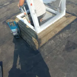 Pracownik wchodzi po drabinie przez otwarty świetlik dachowy na płaskim dachu pokrytym papą, obok stoi niebieska lampa budowlana Makita.