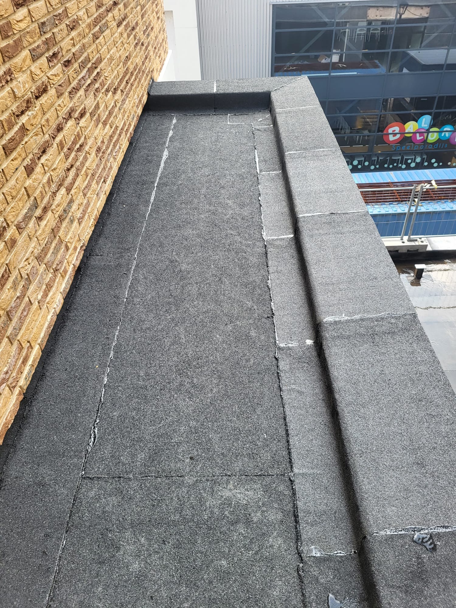 Płaski dach pokryty papą, z widocznymi łączeniami i uszczelnieniami, przylegający do ceglanej ściany budynku. W tle widoczny budynek z dużymi oknami.