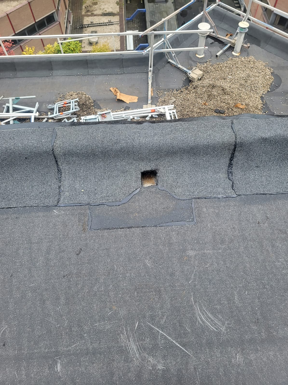 Płaski dach pokryty papą z widocznym wycięciem na wpust dachowy i fragmentami rozsypanego żwiru oraz rusztowaniem w tle.