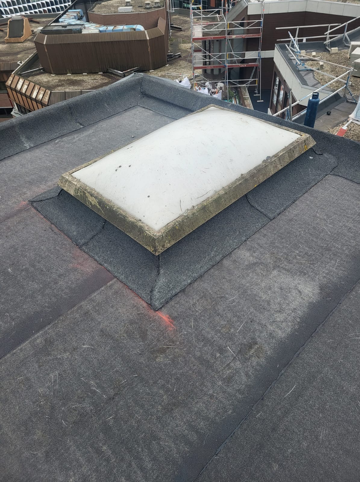 Płaski dach pokryty papą z widocznym świetlikiem dachowym z betonową ramą. W tle inne budynki i rusztowanie.