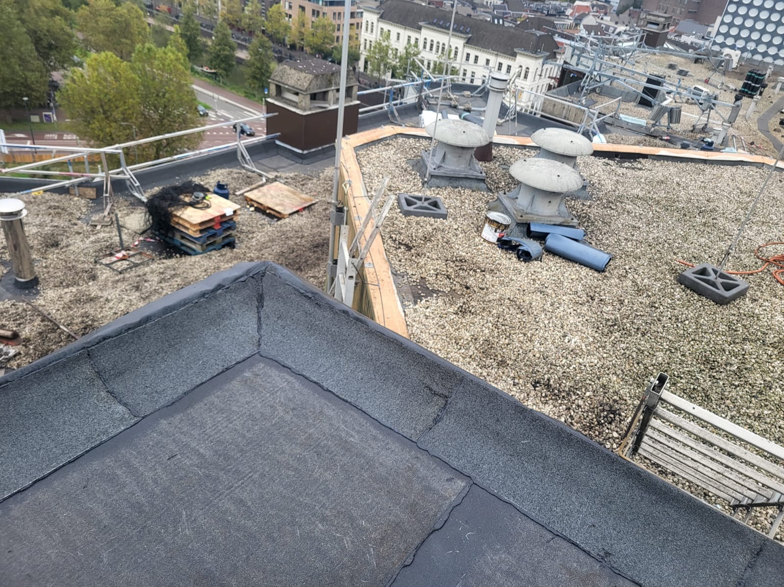 Widok z góry na płaski dach pokryty papą i drobnym żwirem, z elementami wentylacyjnymi, paletami i narzędziami, w tle zabudowa miejska.