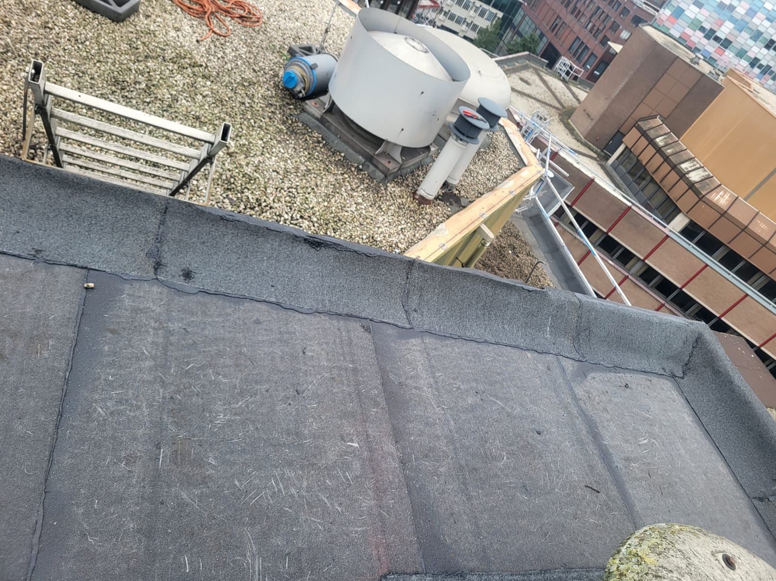 Widok z góry na płaski dach pokryty papą, z widocznymi łączeniami i drobnymi uszkodzeniami, w tle instalacje wentylacyjne, drabina i budynki miejskie.
