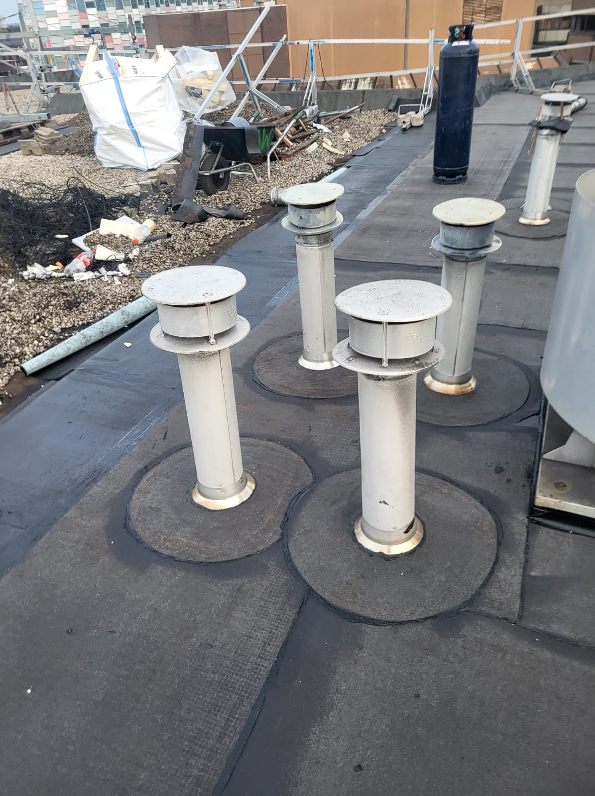 Płaski dach pokryty papą z zamontowanymi kominkami wentylacyjnymi, widoczne ślady prac dekarskich i materiały budowlane w tle.