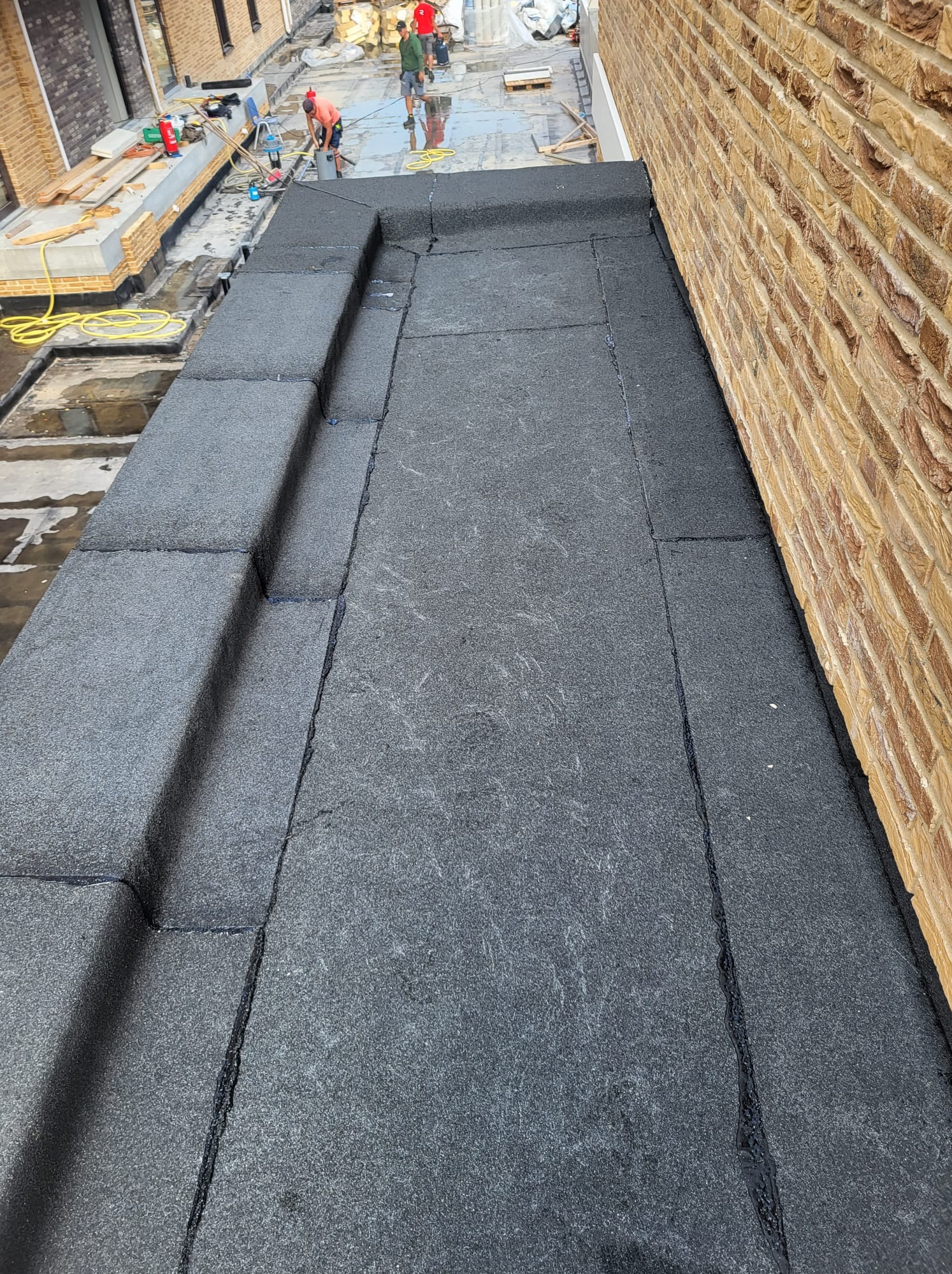 Nowa, płaska powierzchnia dachu pokryta papą termozgrzewalną, z widocznymi pracownikami w tle i żółtym wężem, przylega do ceglanej ściany budynku.