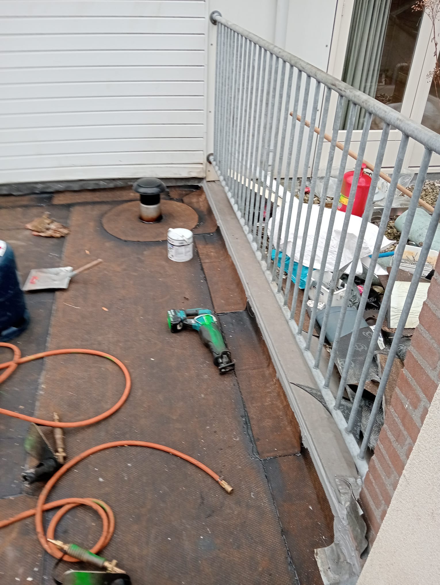 Płaski dach pokryty papą z kominkiem wentylacyjnym, narzędziami i resztkami materiałów budowlanych, zabezpieczony metalową barierką, widok z góry.