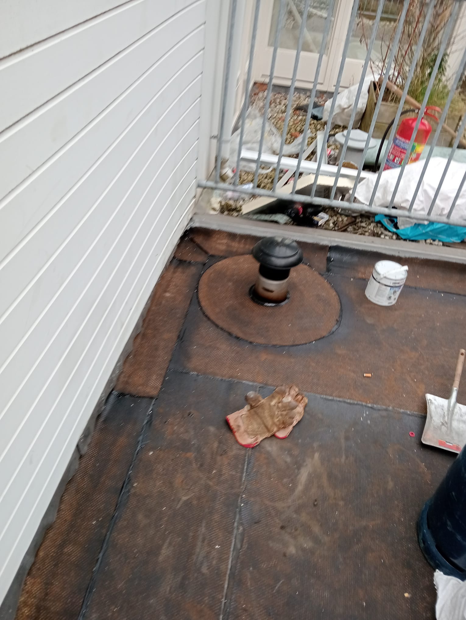 Widok z góry na fragment dachu pokrytego papą, z kominkiem wentylacyjnym, rękawicą roboczą, puszką farby i narzędziem, balkon z metalową barierką w tle.