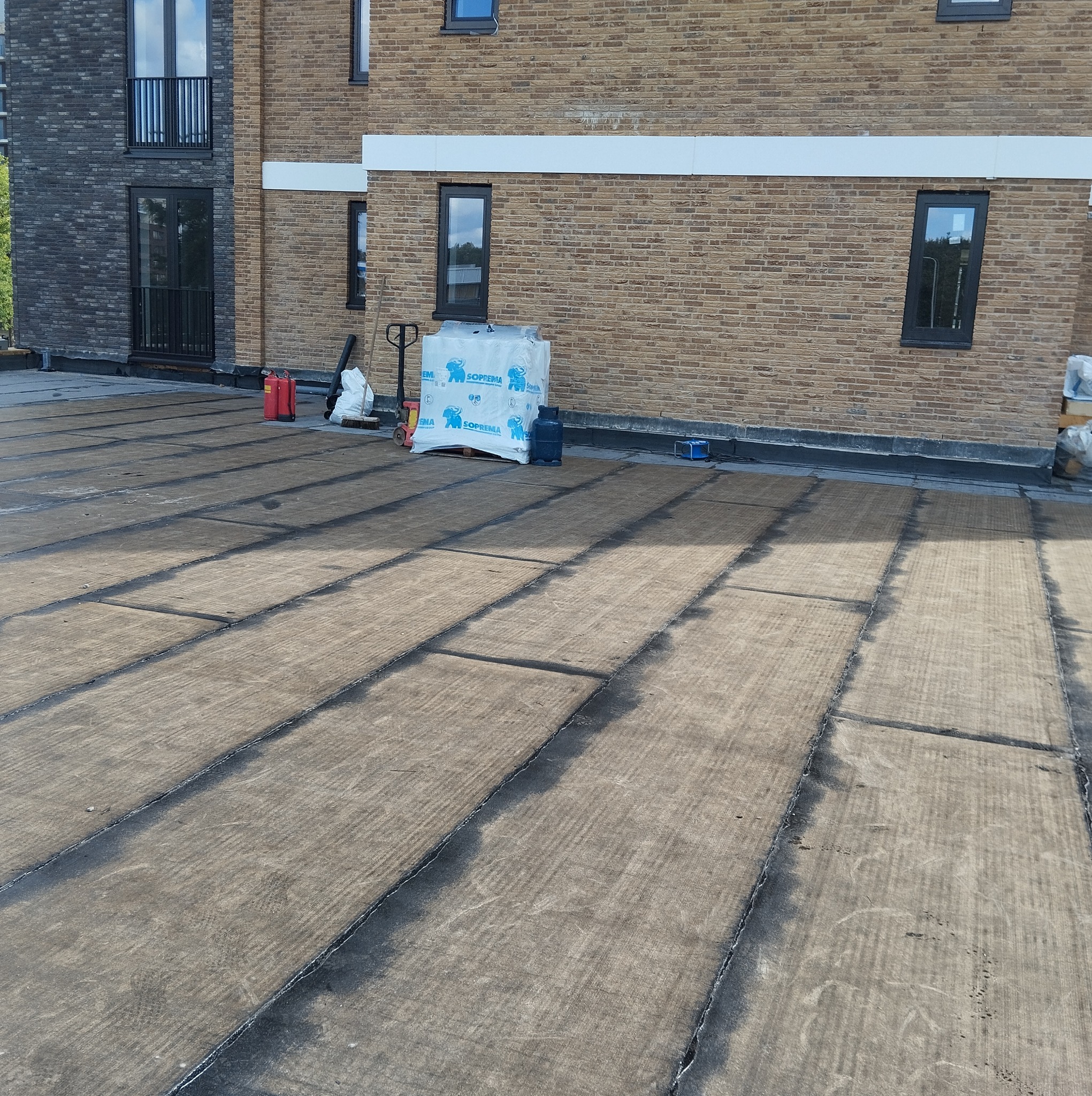 Płaski dach pokryty izolacją termiczną Soprema, przygotowany do dalszych prac, z widocznymi butlami gazowymi i paletą materiałów budowlanych na tle ceglanego budynku.