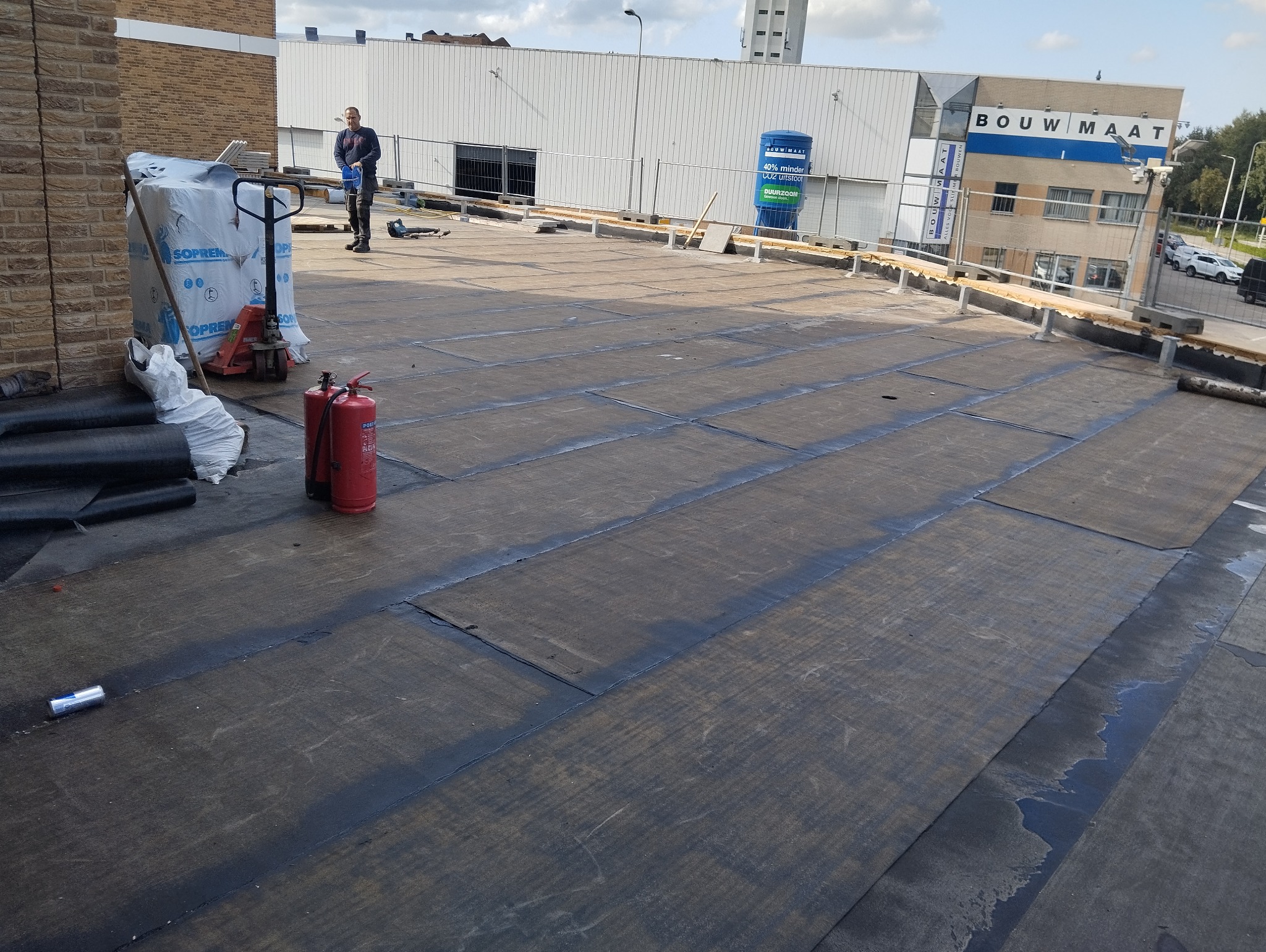 Płaski dach pokryty papą termozgrzewalną, widoczne zgrzewy, butle z gazem, paleta z materiałami budowlanymi i pracownik w tle. Częściowo zakończone prace.