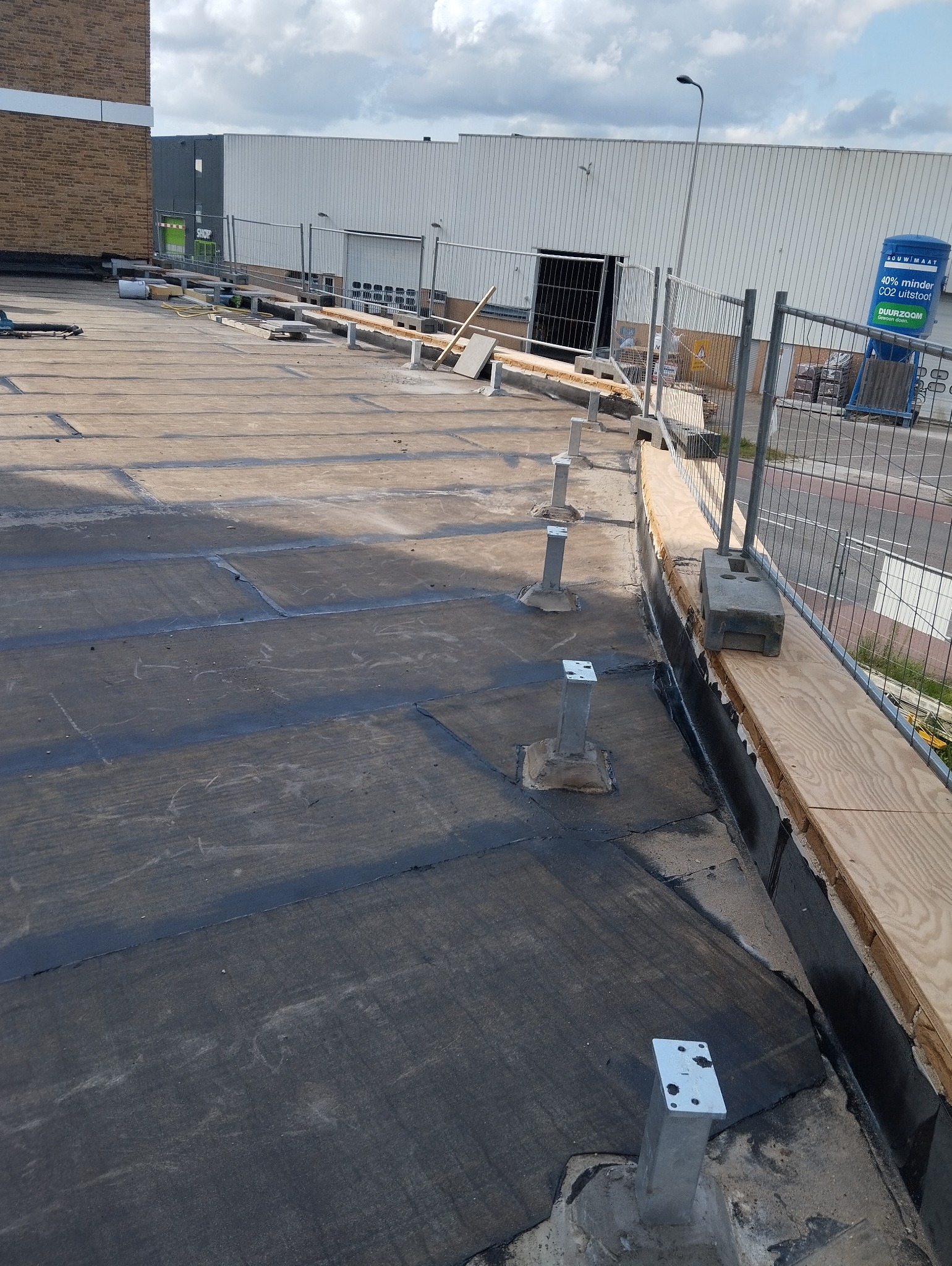 Flat roof under construction with visible bitumen sealing, metal support posts, and temporary wooden edge protection near a building.