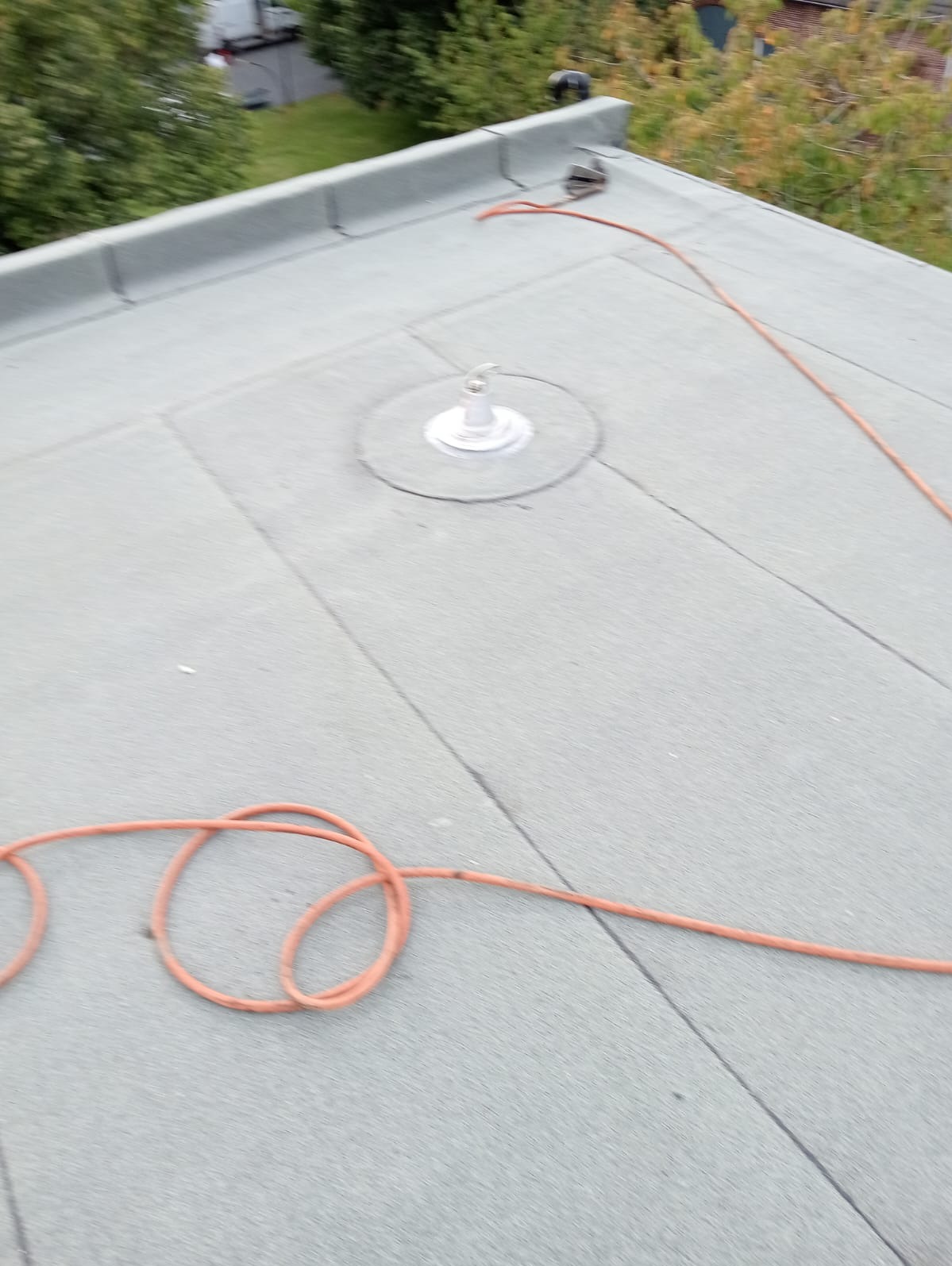 Płaski dach pokryty papą z widocznym kominkiem wentylacyjnym i pomarańczowym przewodem na powierzchni.