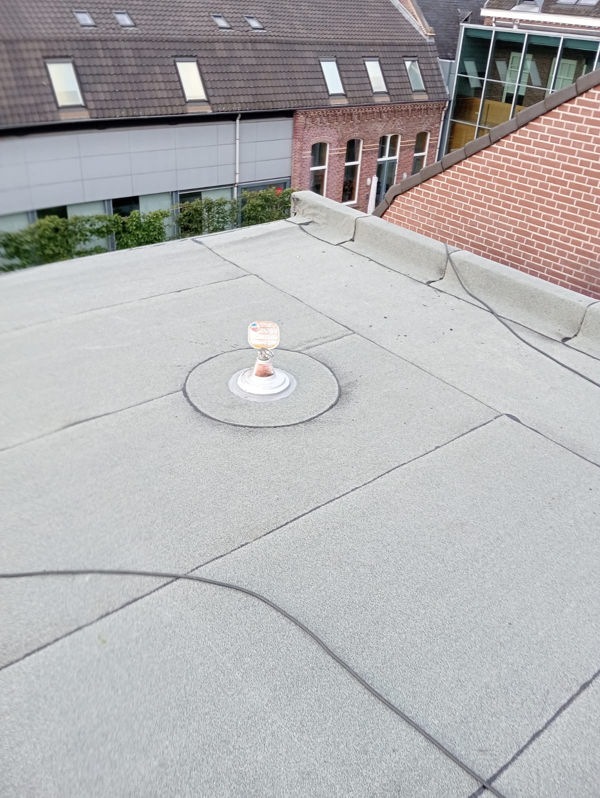 Płaski dach pokryty papą z widocznym kominkiem wentylacyjnym i szwem, na tle budynki z cegły i szarej blachy