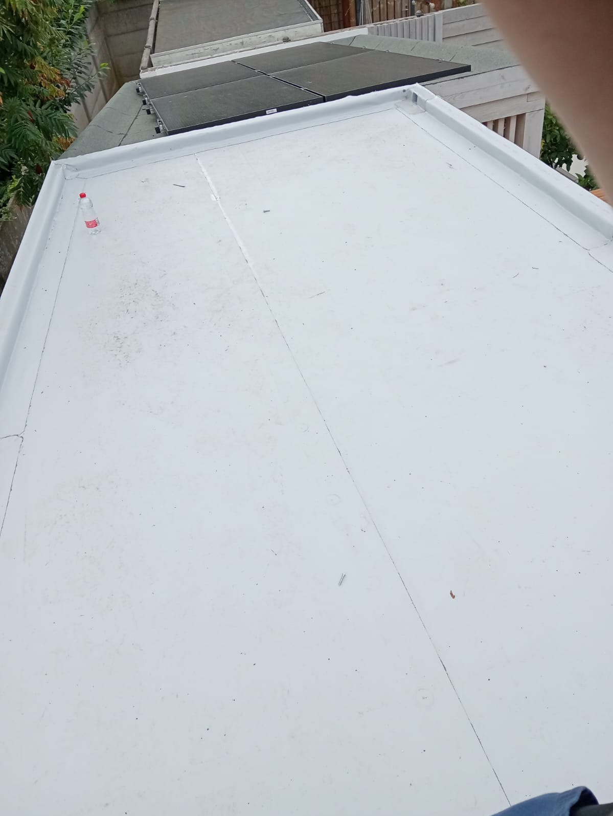 Biały, płaski dach pokryty papą, z widocznymi panelami solarnymi w tle i butelką wody na powierzchni.