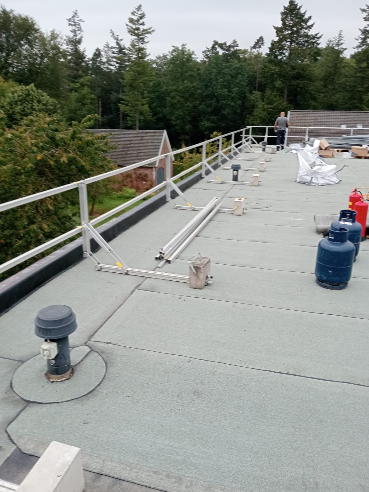 Płaski dach pokryty papą termozgrzewalną z zamontowaną aluminiową barierką ochronną, widoczne butle gazowe, elementy wentylacyjne i pracownik w oddali.