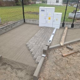 KWS - Fragment układania szarej kostki brukowej na podsypce piaskowej, widoczny poziomica i krawężnik, w tle ogrodzenie panelowe i skrzynki elektryczne.