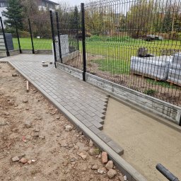 KWS - Układanie szarej kostki brukowej przy ogrodzeniu panelowym z podmurówką betonową, widoczne narzędzia i przygotowane podłoże.