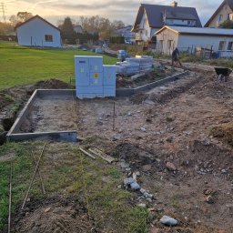 KWS - Przygotowanie terenu pod kostkę brukową: wykopany fundament z betonową obwódką, widoczny elektryczny panel sterujący, ułożone obok kostki, narzędzia i pracownik z taczką w tle.