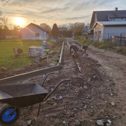 KWS - Prace przygotowawcze do ułożenia nawierzchni z kostki brukowej w trakcie zachodu słońca, widoczny pracownik z narzędziami, obrzeża betonowe, paleta kostki brukowej oraz taczki.