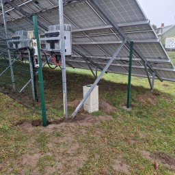 KWS - Widok od spodu na panele słoneczne i fragment ogrodzenia z siatki, z poziomicą przytwierdzoną do słupka, w Sławnie, na ziemi widoczne świeże ślady po montażu.