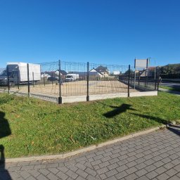 KWS - Ciemnoszara siatka ogrodzeniowa na betonowej podmurówce, otaczająca teren z piaskiem; w tle zaparkowany biały samochód dostawczy i zabudowa jednorodzinna w słoneczny dzień.