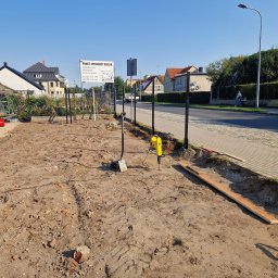 KWS - Przygotowany teren pod montaż ogrodzenia z czarnymi słupkami; widoczny żółty młot udarowy wbijający słupek, ziemia z gruzem, w tle chodnik i zabudowania.
