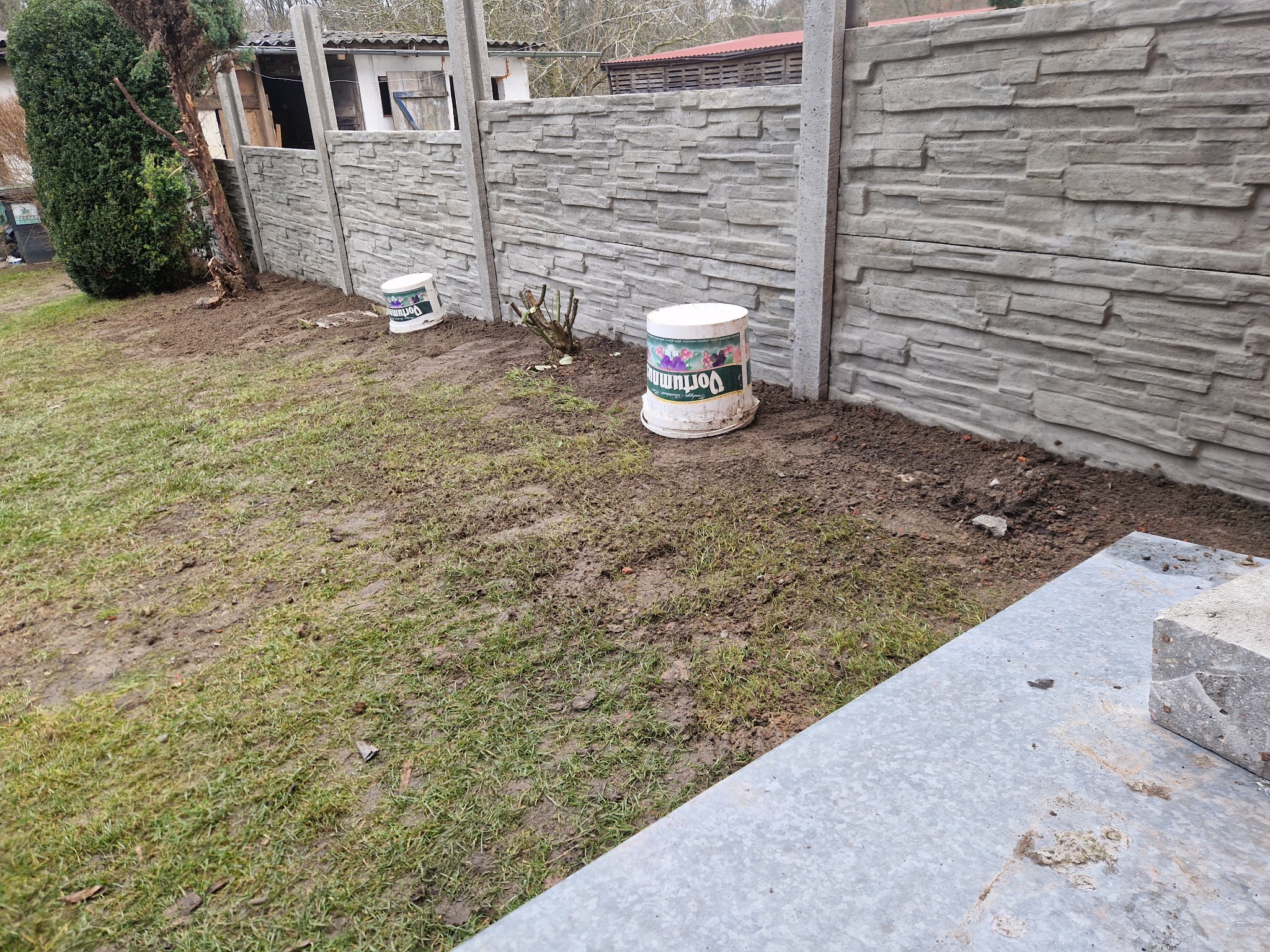 Nowo postawione ogrodzenie betonowe imitujące kamień, z dwoma wiaderkami po farbie u podstawy, na trawniku z widocznymi śladami prac ziemnych.