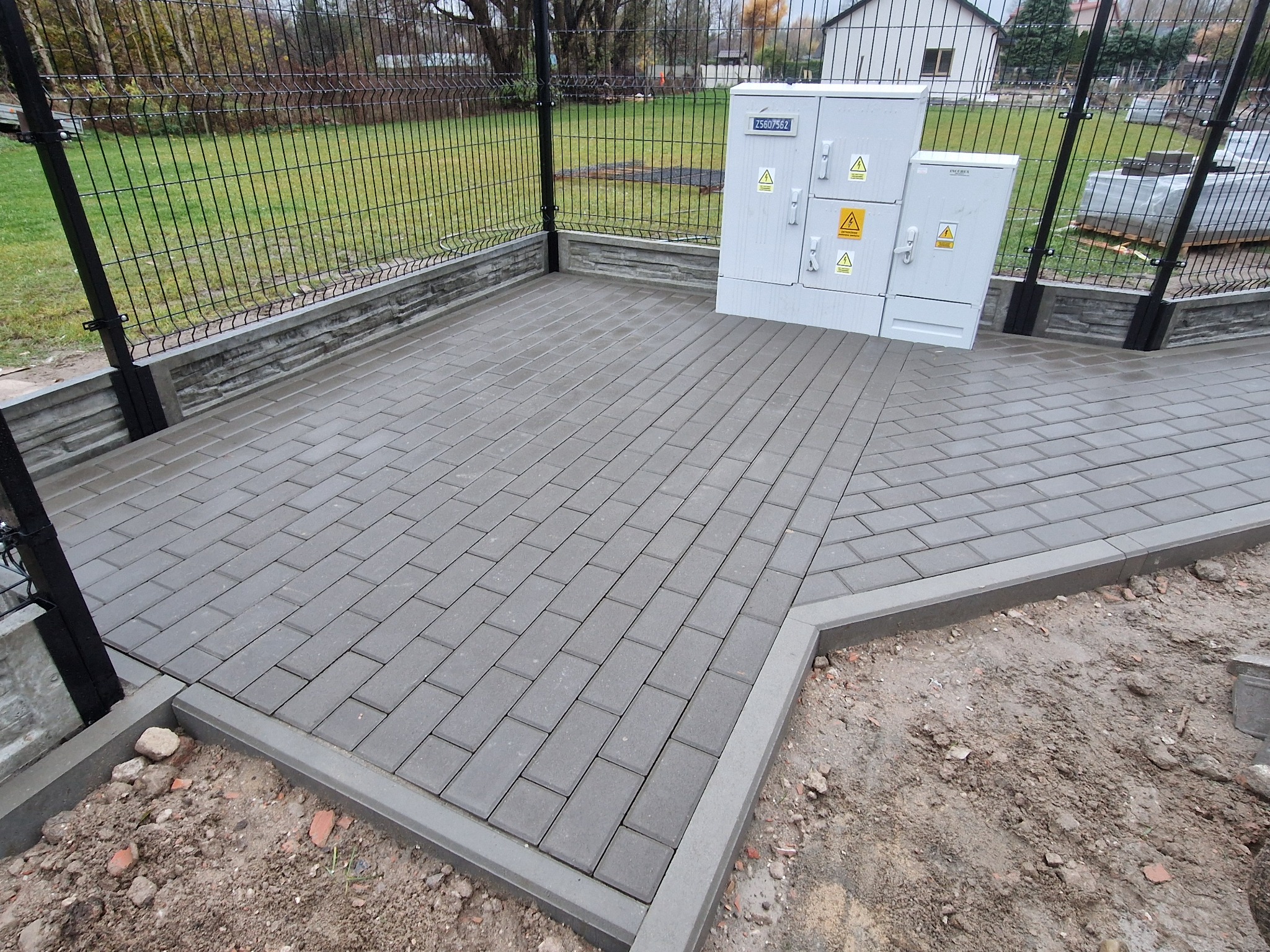 Ułożona szara kostka brukowa na podwórku, obok ogrodzenia z siatki i skrzynki elektrycznej, widoczne fragmenty ziemi i trawy.