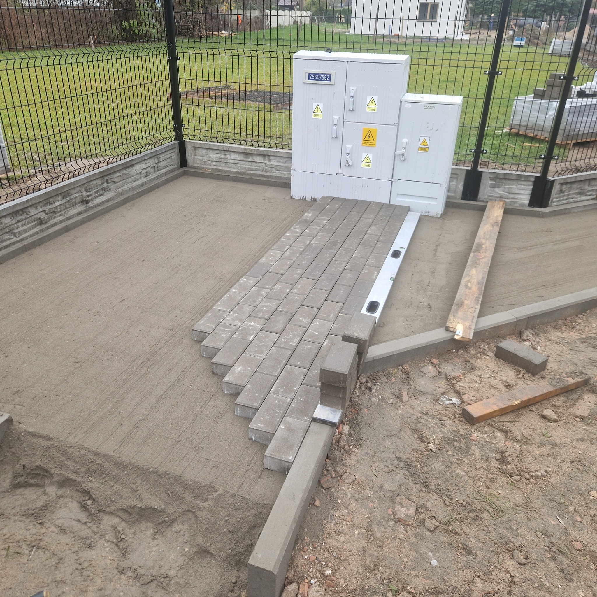 Fragment układania szarej kostki brukowej na podsypce piaskowej, widoczny poziomica i krawężnik, w tle ogrodzenie panelowe i skrzynki elektryczne.