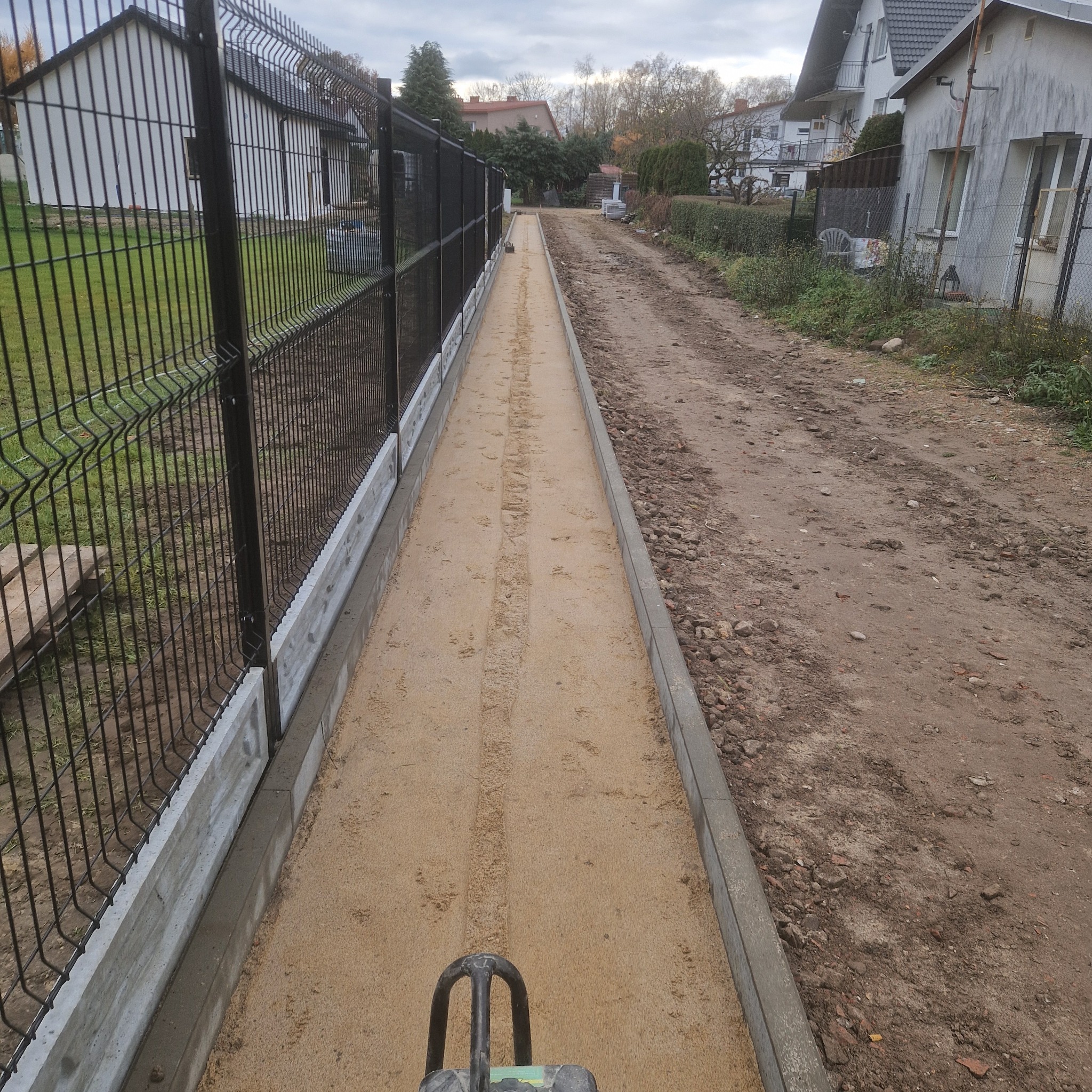 Przygotowanie podłoża piaskiem pod ścieżkę z obrzeżami betonowymi i ogrodzeniem panelowym, widoczne ślady ubijarki.