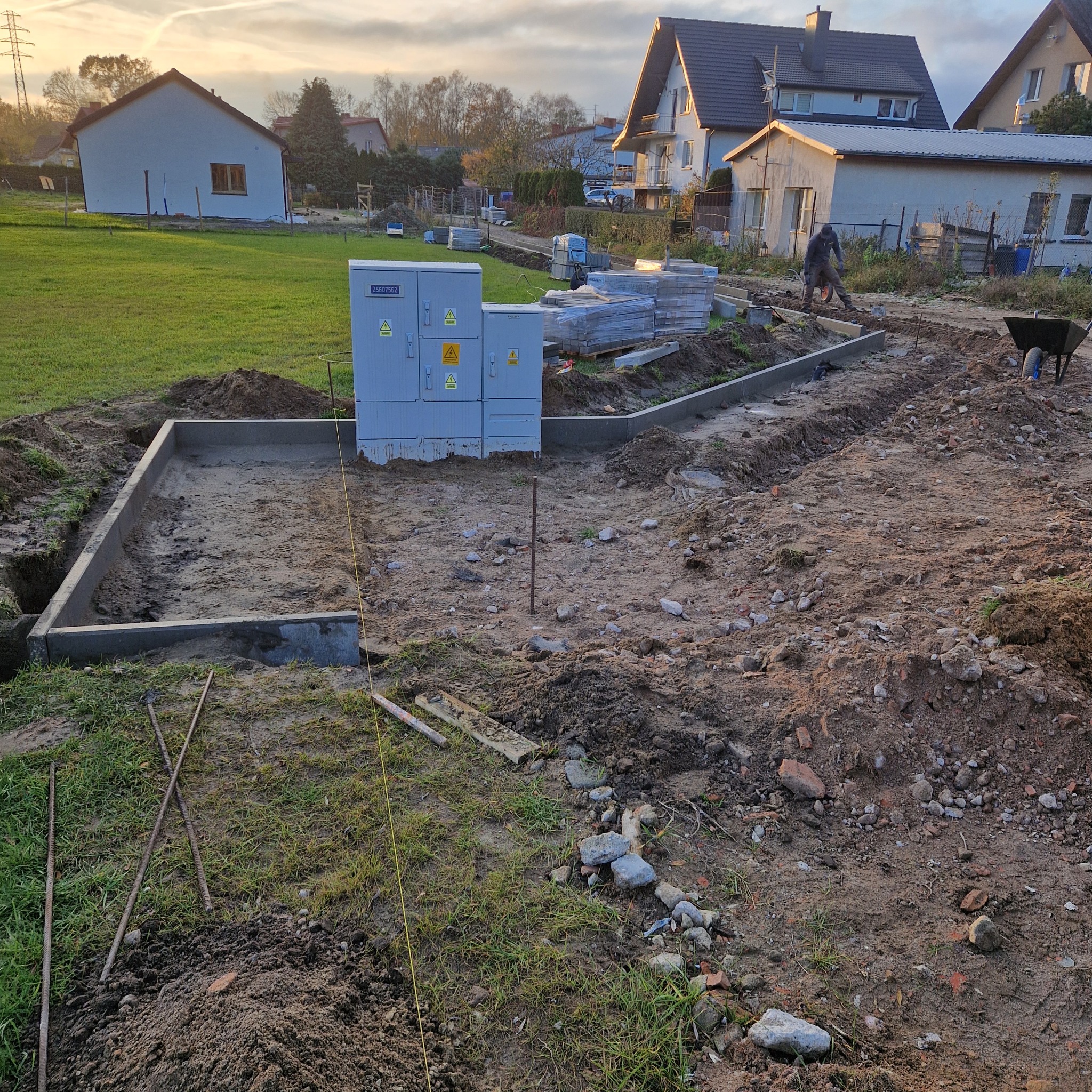 Przygotowanie terenu pod kostkę brukową: wykopany fundament z betonową obwódką, widoczny elektryczny panel sterujący, ułożone obok kostki, narzędzia i pracownik z taczką w tle.