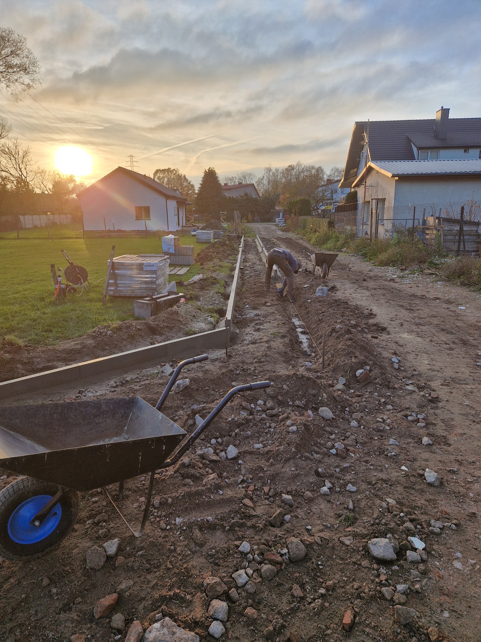 Prace przygotowawcze do ułożenia nawierzchni z kostki brukowej w trakcie zachodu słońca, widoczny pracownik z narzędziami, obrzeża betonowe, paleta kostki brukowej oraz taczki.