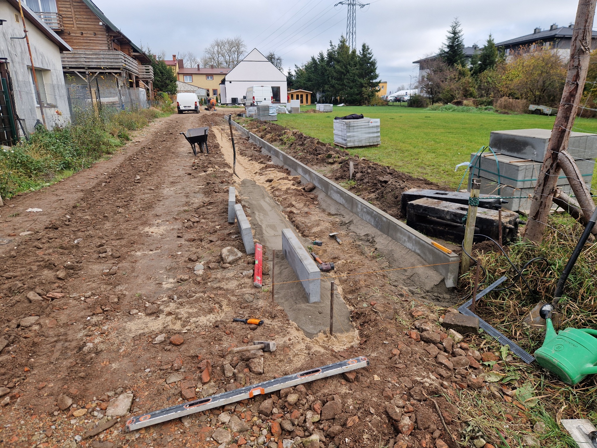 Przygotowanie podłoża pod chodnik z kostki brukowej: widoczne krawężniki, poziomice, sznurek wyznaczający linię, taczka w tle, teren częściowo wyłożony gruzem ceglanym.