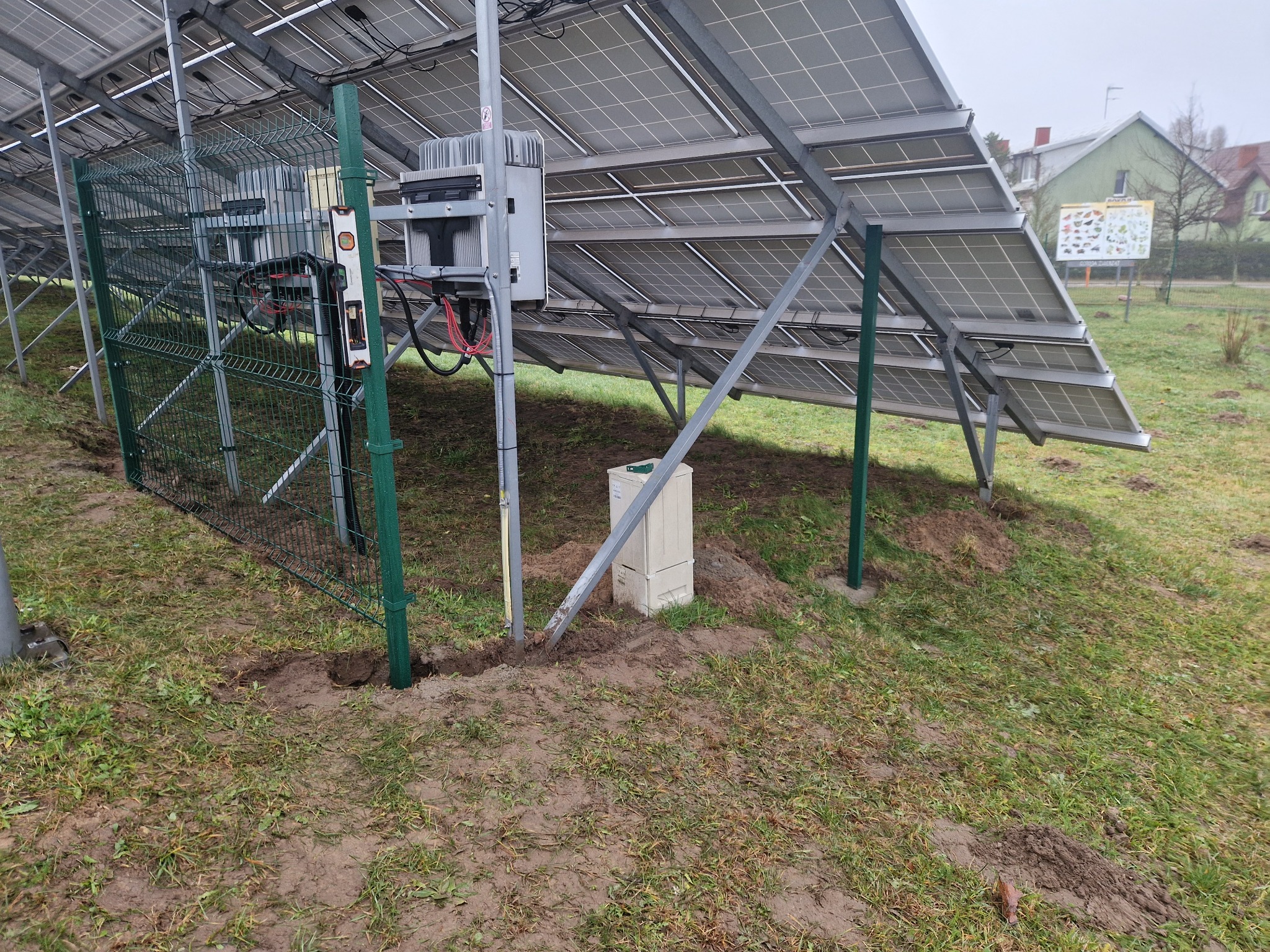 Widok od spodu na panele słoneczne i fragment ogrodzenia z siatki, z poziomicą przytwierdzoną do słupka, w Sławnie, na ziemi widoczne świeże ślady po montażu.