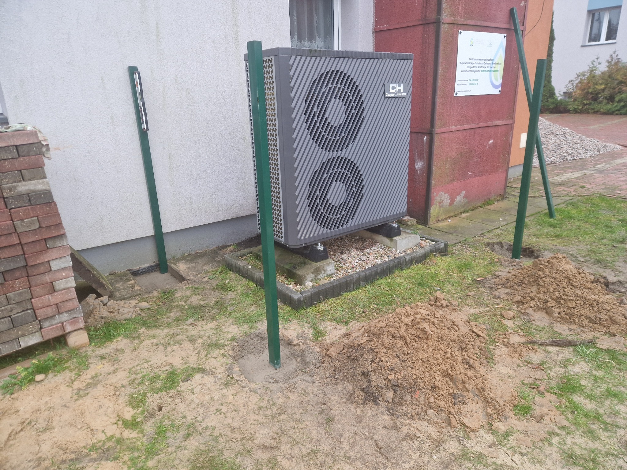 Przygotowania do montażu ogrodzenia: wbite słupki w ziemię obok ułożonej kostki brukowej pod pompę ciepła Cooper&Hunter, sterta kostki z boku.