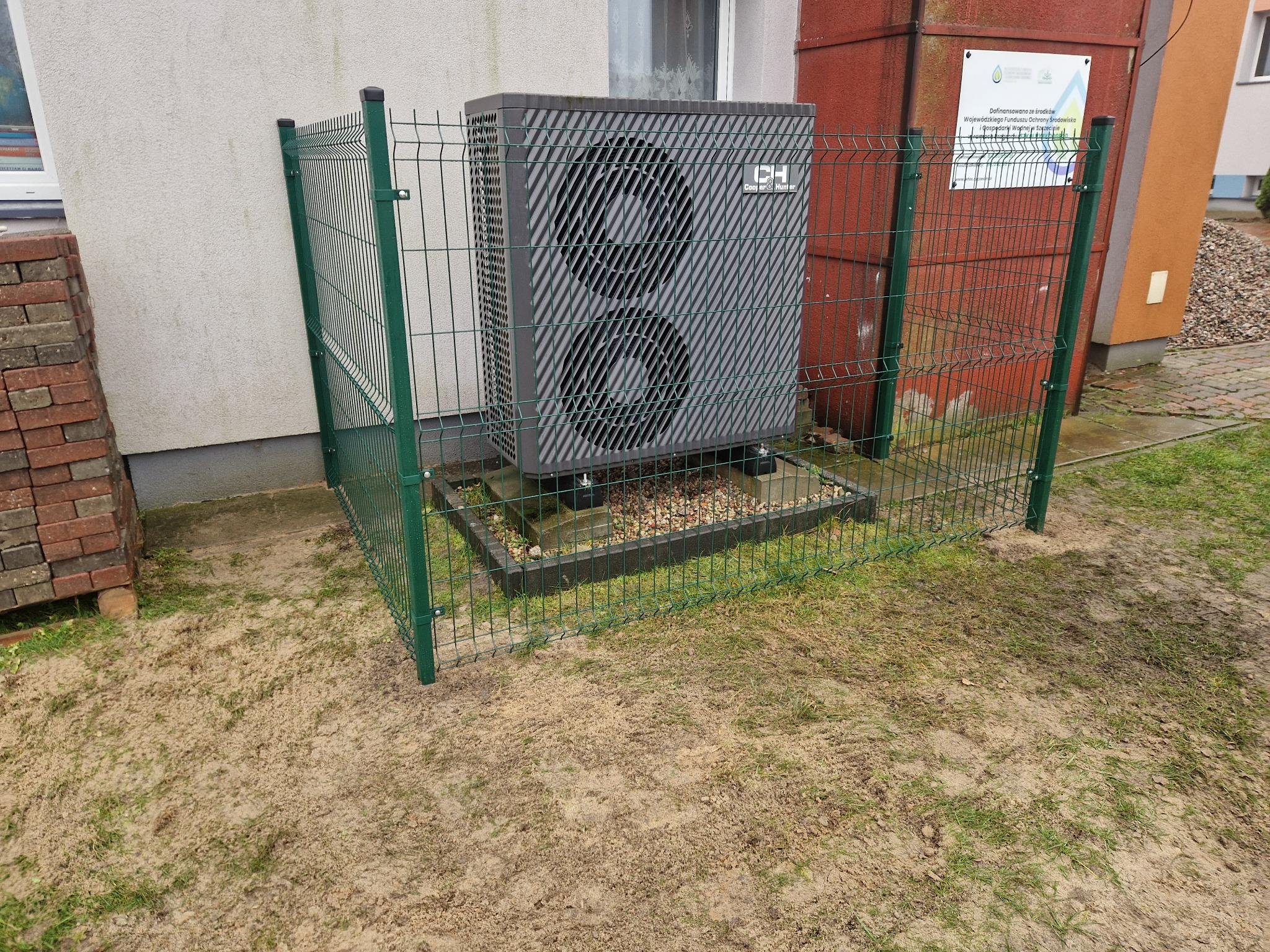 Szara pompa ciepła marki Cooper&Hunter, osłonięta zielonym ogrodzeniem panelowym na podwórku z piaskiem i trawą, obok budynku z cegłami i tablicą informacyjną.
