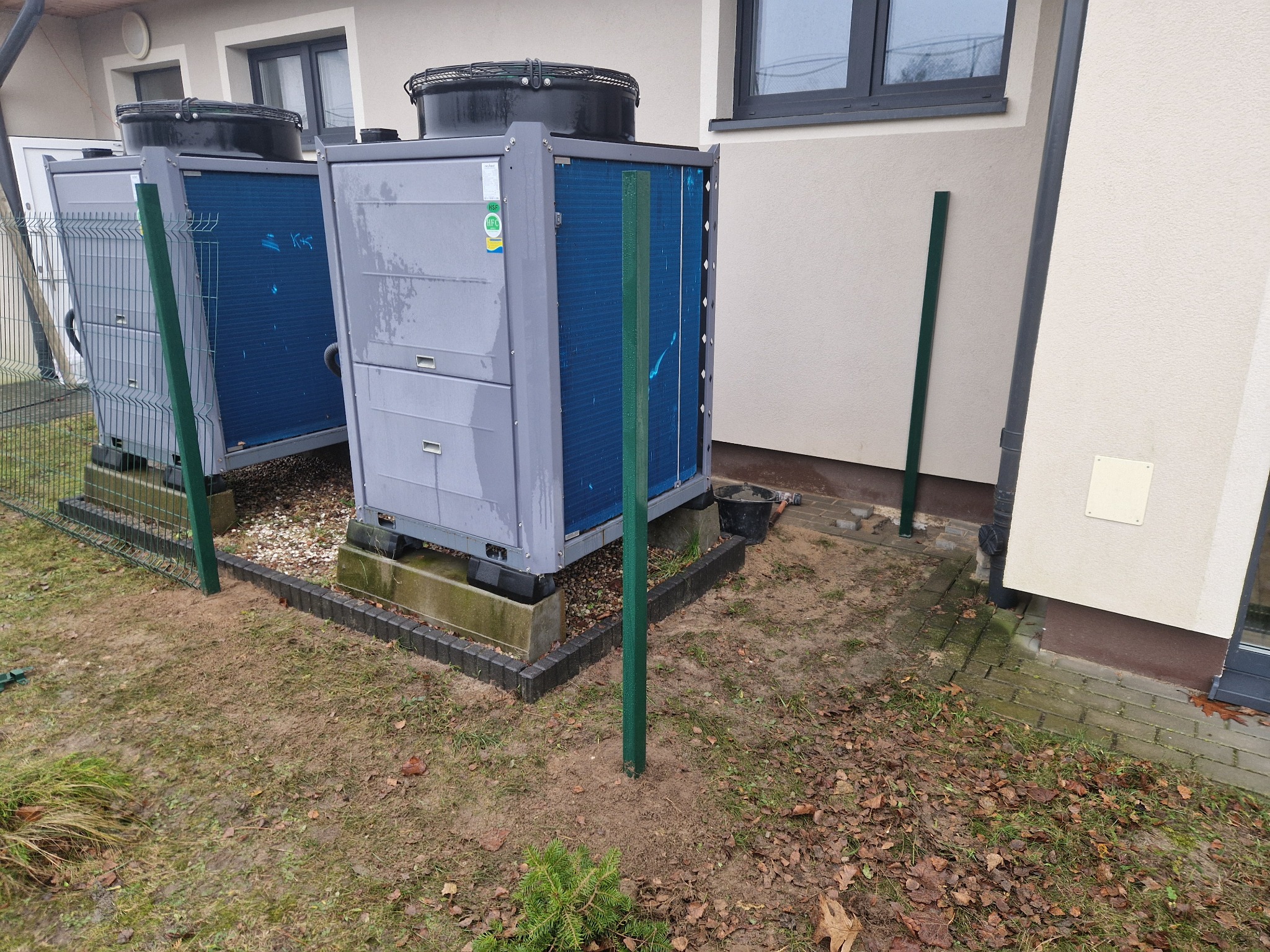 Dwie szare pompy ciepła ustawione na betonowych podstawach obok budynku z jasną elewacją. Wokół widać przygotowany teren z wbitymi słupkami ogrodzeniowymi i resztkami liści.