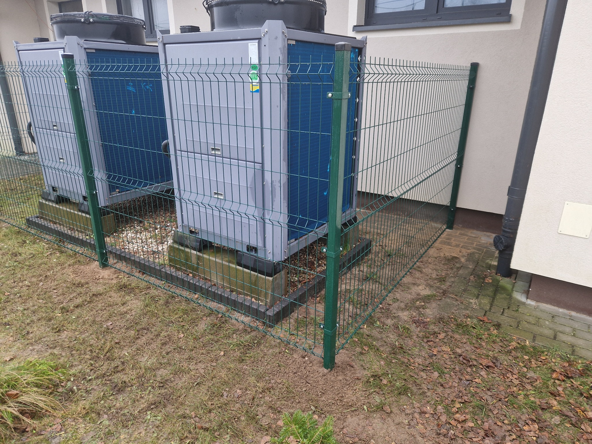 Ochrona agregatów klimatyzacyjnych ogrodzeniem panelowym z zielonymi słupkami na trawiastym podłożu.